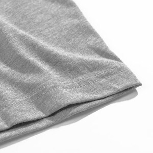 Pu&ograve; includere: Primo piano dell'orlo di una maglietta grigia. Il tessuto &egrave; un materiale morbido a maglia con cuciture visibili lungo il bordo. La maglietta &egrave; di colore grigio chiaro.