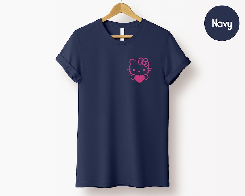 May include: Navy blue t-shirt featuring a pink Hello Kitty graphic on the chest. The graphic includes the character's face, bow, and a heart. The shirt has short sleeves and a crew neck.