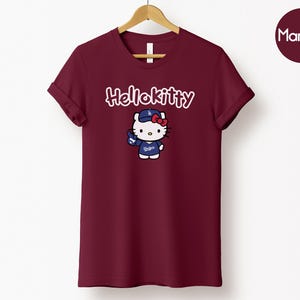May include: Maroon t-shirt with a Hello Kitty graphic. The graphic depicts Hello Kitty in a baseball cap and uniform. The text "Hello Kitty" is above the graphic in cursive. The word "Maroon" is in a circle in the top right corner.