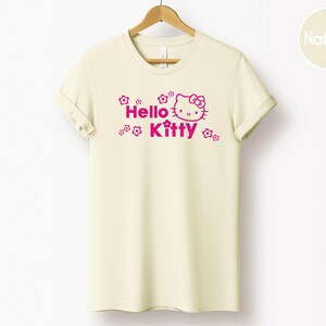May include: A cream-coloured t-shirt featuring the words "Hello Kitty" in pink, accompanied by a graphic of Hello Kitty's face and small flower designs. The shirt is displayed on a wooden hanger.