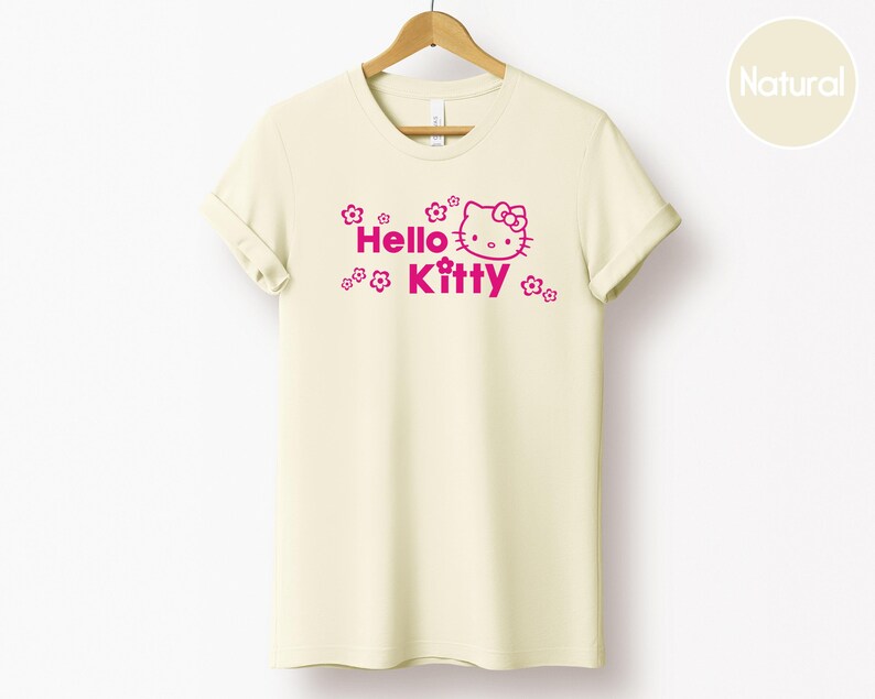 May include: A cream-coloured t-shirt featuring the words "Hello Kitty" in pink, accompanied by a graphic of Hello Kitty's face and small flower designs. The shirt is displayed on a wooden hanger.