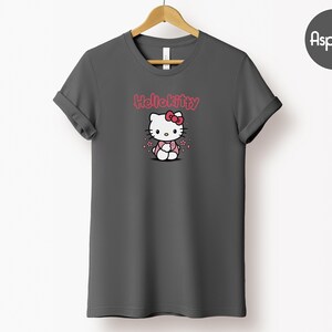 May include: A dark gray t-shirt featuring a Hello Kitty graphic. The graphic includes the text "Hello Kitty" in pink above an illustration of the character wearing a pink bow and outfit. The shirt is hanging on a wooden hanger.