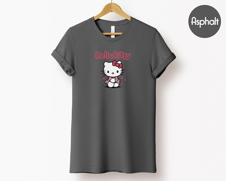 May include: A dark gray t-shirt featuring a Hello Kitty graphic. The graphic includes the text "Hello Kitty" in pink above an illustration of the character wearing a pink bow and outfit. The shirt is hanging on a wooden hanger.