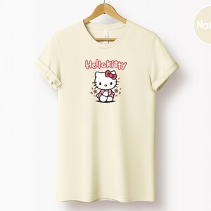 May include: Cream-colored t-shirt featuring a Hello Kitty graphic. The design includes the text "Hello Kitty" in pink above an illustration of the character with a pink bow and flowers. The shirt is hanging on a wooden hanger.