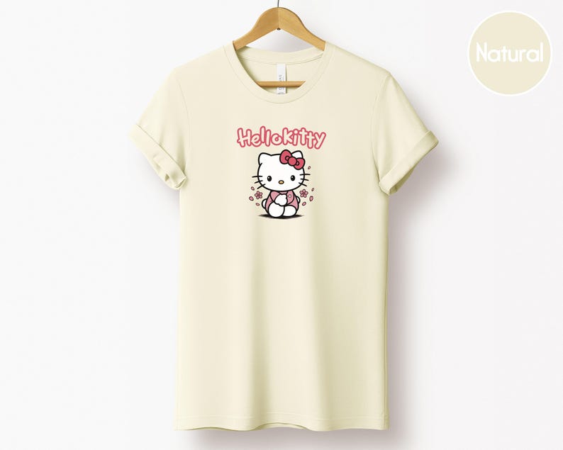 May include: Cream-colored t-shirt featuring a Hello Kitty graphic. The design includes the text "Hello Kitty" in pink above an illustration of the character with a pink bow and flowers. The shirt is hanging on a wooden hanger.