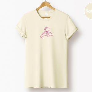May include: A cream-colored t-shirt with a pink graphic of a cartoon cat riding a dolphin. The cat is wearing a bow and the dolphin is smiling. The shirt is hanging on a wooden hanger.