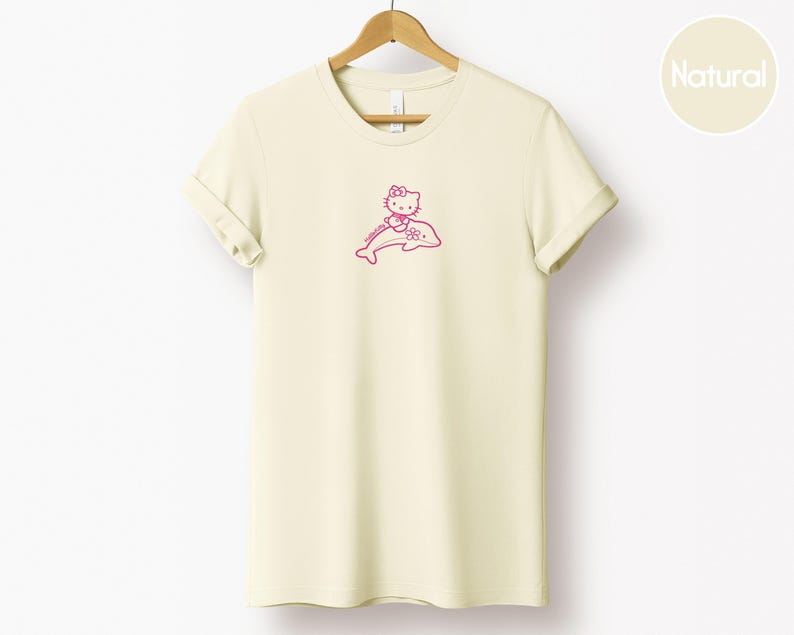 May include: A cream-colored t-shirt with a pink graphic of a cartoon cat riding a dolphin. The cat is wearing a bow and the dolphin is smiling. The shirt is hanging on a wooden hanger.