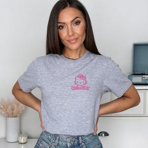 May include: A heather gray t-shirt featuring a pink Hello Kitty graphic. The graphic includes a bow and the text "HelloKitty". The shirt is short-sleeved and made of a soft material.
