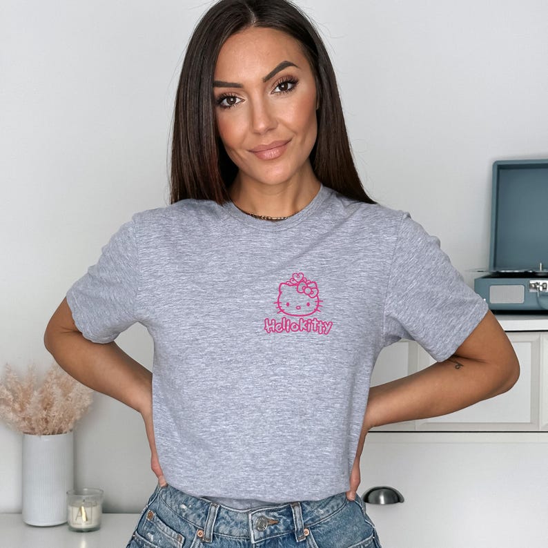 May include: A heather gray t-shirt featuring a pink Hello Kitty graphic. The graphic includes a bow and the text "HelloKitty". The shirt is short-sleeved and made of a soft material.