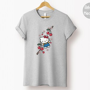 Op de afbeelding: Grijze t-shirt met een cartoonfiguur, een rode strik, blauwe overalls en een kersenboomtak. Het t-shirt hangt aan een houten hanger. De tekst "Athletic Heather" staat in een cirkel rechts.