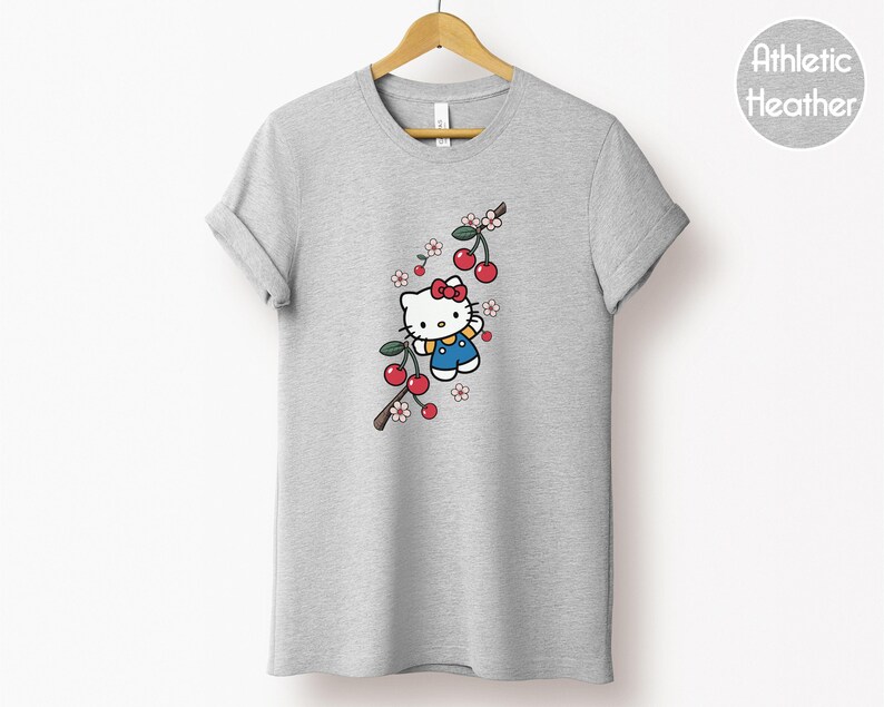 Op de afbeelding: Grijze t-shirt met een cartoonfiguur, een rode strik, blauwe overalls en een kersenboomtak. Het t-shirt hangt aan een houten hanger. De tekst "Athletic Heather" staat in een cirkel rechts.