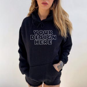Peut inclure: Un sweat à capuche noir avec une poche avant et le texte "YOUR DESIGN HERE" en lettres blanches. Le sweat à capuche a une capuche à cordon et est porté par une personne.