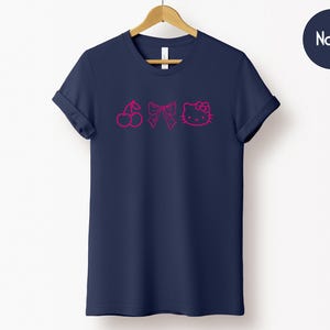 Pu&ograve; includere: T-shirt blu navy con grafica rosa a ciliegia, fiocco e faccina di gatto. La t-shirt &egrave; esposta su una gruccia di legno su sfondo bianco. La parola "Navy" &egrave; in un cerchio blu navy in alto a destra.
