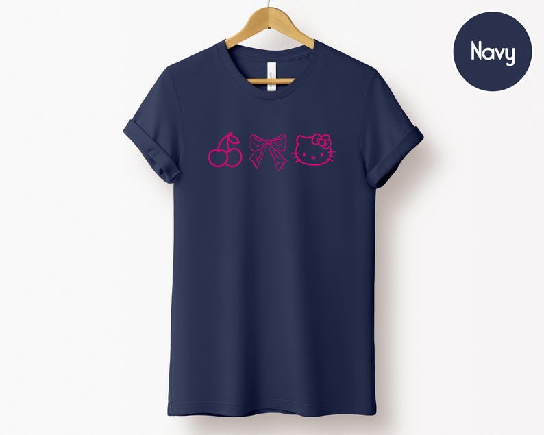 Pu&ograve; includere: T-shirt blu navy con grafica rosa a ciliegia, fiocco e faccina di gatto. La t-shirt &egrave; esposta su una gruccia di legno su sfondo bianco. La parola "Navy" &egrave; in un cerchio blu navy in alto a destra.