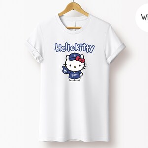 May include: White t-shirt with a Hello Kitty graphic in a baseball uniform. The design includes the text "Hello Kitty" in blue, and Hello Kitty is wearing a blue cap and uniform with the Dodgers logo. A red bow completes the look.
