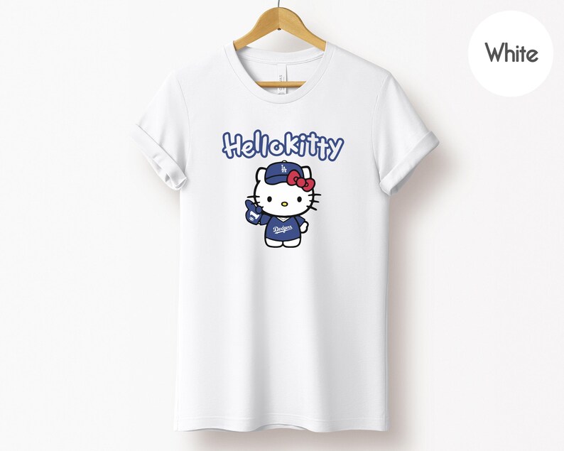 May include: White t-shirt with a Hello Kitty graphic in a baseball uniform. The design includes the text "Hello Kitty" in blue, and Hello Kitty is wearing a blue cap and uniform with the Dodgers logo. A red bow completes the look.