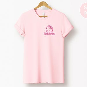 May include: A light pink t-shirt with a Hello Kitty graphic in the upper left chest area. The graphic includes the Hello Kitty character and the text "HelloKitty" in a matching pink color. The t-shirt is hanging on a wooden hanger.