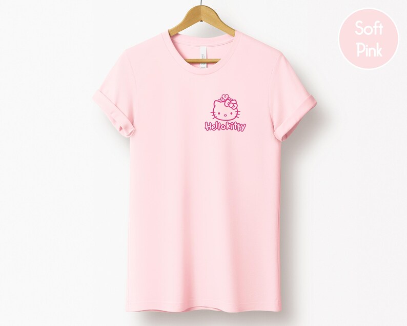 May include: A light pink t-shirt with a Hello Kitty graphic in the upper left chest area. The graphic includes the Hello Kitty character and the text "HelloKitty" in a matching pink color. The t-shirt is hanging on a wooden hanger.