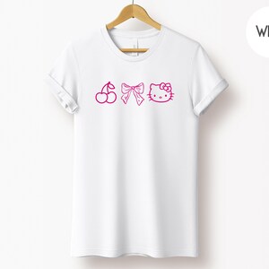 Pu&ograve; includere: T-shirt bianca con illustrazioni rosa di ciliegie, un fiocco e Hello Kitty. La maglietta &egrave; appesa a un appendiabiti di legno. Un cerchio bianco con la scritta "White" si trova nell'angolo in alto a destra.
