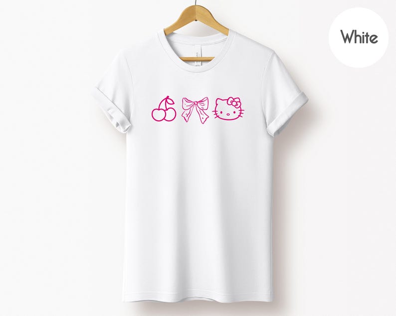 Pu&ograve; includere: T-shirt bianca con illustrazioni rosa di ciliegie, un fiocco e Hello Kitty. La maglietta &egrave; appesa a un appendiabiti di legno. Un cerchio bianco con la scritta "White" si trova nell'angolo in alto a destra.