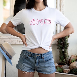 Pu&ograve; includere: T-shirt bianco con grafica rosa raffigurante una ciliegia, un fiocco e una faccia di gatto. La maglietta &egrave; abbinata a pantaloncini di jeans. Un cappello di paglia con tesa blu &egrave; visibile sullo sfondo.