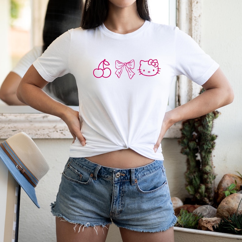 Pu&ograve; includere: T-shirt bianco con grafica rosa raffigurante una ciliegia, un fiocco e una faccia di gatto. La maglietta &egrave; abbinata a pantaloncini di jeans. Un cappello di paglia con tesa blu &egrave; visibile sullo sfondo.