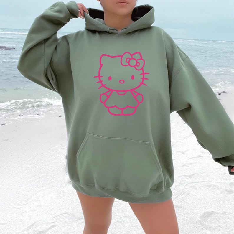 May include: An olive green hoodie with a pink graphic of a cartoon cat character. The hoodie has a kangaroo pocket and a drawstring hood. The image is taken outdoors, with a beach and ocean in the background.