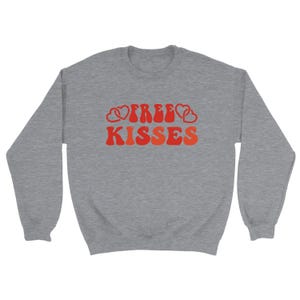 May include: A grey sweatshirt featuring the text "FREE KISSES" in a retro style. The lettering is in shades of red and orange, with heart shapes integrated into the design. The sweatshirt has a crew neck and long sleeves.