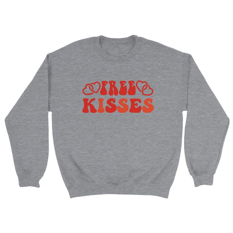 May include: A grey sweatshirt featuring the text "FREE KISSES" in a retro style. The lettering is in shades of red and orange, with heart shapes integrated into the design. The sweatshirt has a crew neck and long sleeves.