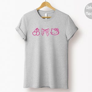 Pu&ograve; includere: Una maglietta grigio melange con un design rosa di ciliegia, fiocco e Hello Kitty. La maglietta &egrave; appesa a una gruccia di legno su sfondo bianco. Il testo "Athletic Heather" &egrave; in un cerchio in alto a destra.