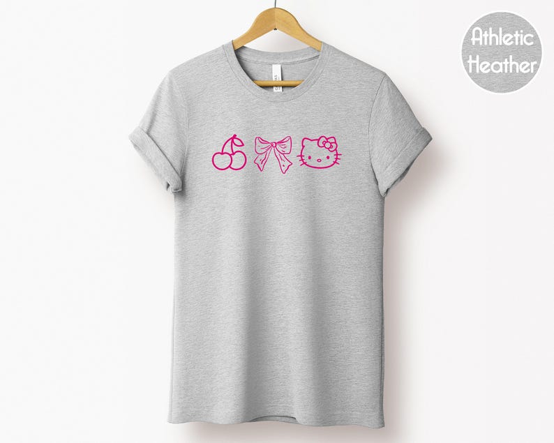 Pu&ograve; includere: Una maglietta grigio melange con un design rosa di ciliegia, fiocco e Hello Kitty. La maglietta &egrave; appesa a una gruccia di legno su sfondo bianco. Il testo "Athletic Heather" &egrave; in un cerchio in alto a destra.