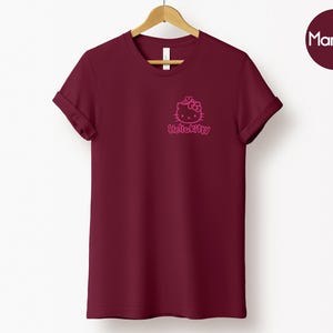 May include: Maroon t-shirt with a pink Hello Kitty graphic on the chest. The short-sleeved shirt is hanging on a wooden hanger against a white background. A maroon circle with the word "Maroon" is in the upper right corner.
