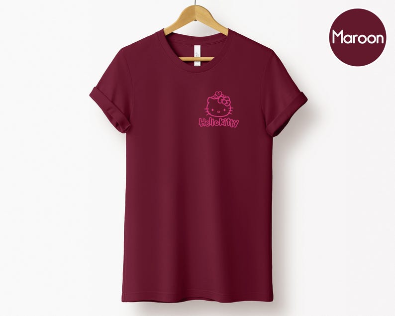 May include: Maroon t-shirt with a pink Hello Kitty graphic on the chest. The short-sleeved shirt is hanging on a wooden hanger against a white background. A maroon circle with the word "Maroon" is in the upper right corner.