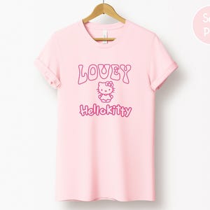 Op de afbeelding: Een lichtroze t-shirt met de tekst "LOVEY" boven een Hello Kitty-afbeelding en het woord "Hellokitty" in roze. Het t-shirt hangt aan een houten hanger.