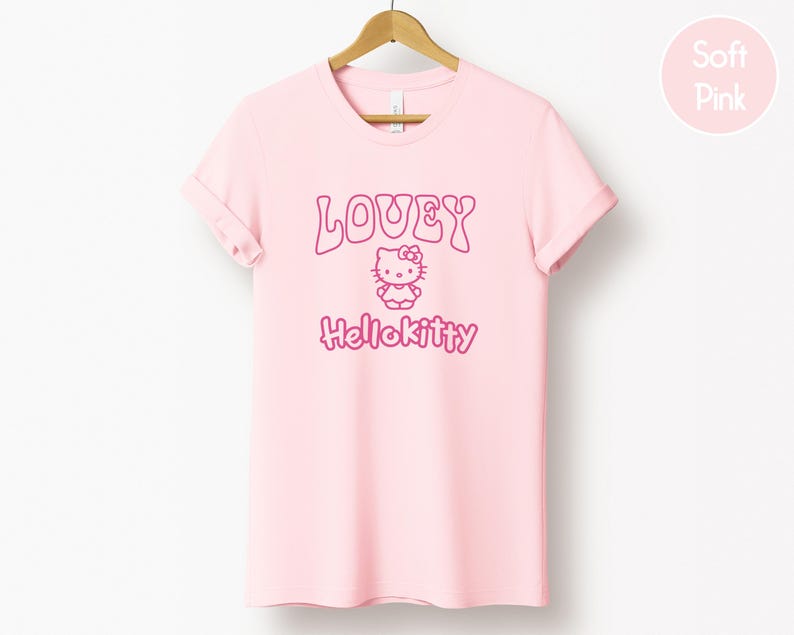 Op de afbeelding: Een lichtroze t-shirt met de tekst "LOVEY" boven een Hello Kitty-afbeelding en het woord "Hellokitty" in roze. Het t-shirt hangt aan een houten hanger.