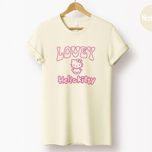 Op de afbeelding: Cr&egrave;mekleurig T-shirt met de roze tekst "LOVEY" en "Hellokitty", met een Hello Kitty-afbeelding. Het t-shirt hangt aan een houten hanger tegen een witte achtergrond.