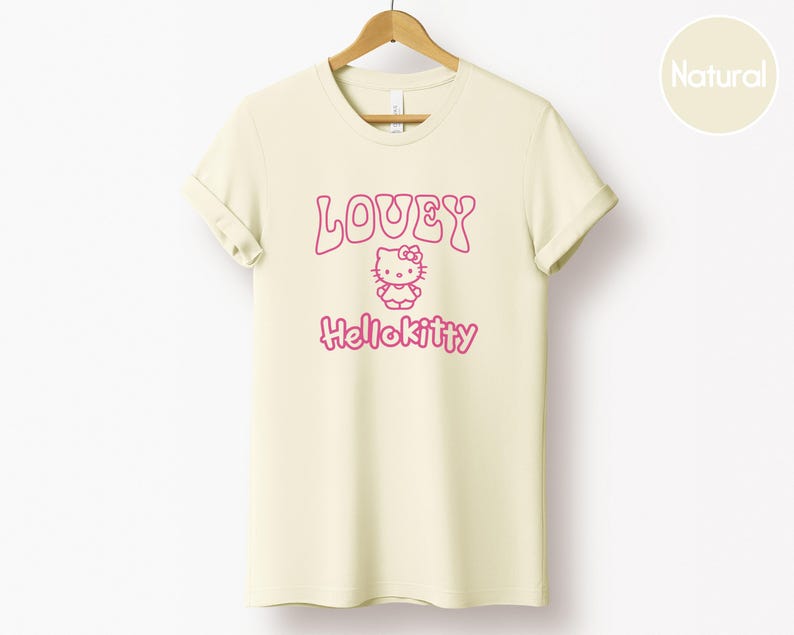 Op de afbeelding: Cr&egrave;mekleurig T-shirt met de roze tekst "LOVEY" en "Hellokitty", met een Hello Kitty-afbeelding. Het t-shirt hangt aan een houten hanger tegen een witte achtergrond.