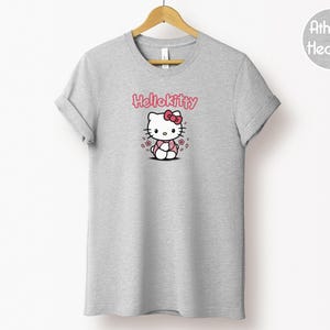 May include: A light gray t-shirt with a graphic of Hello Kitty. The design features Hello Kitty with a pink bow, surrounded by pink flowers, and the text "Hello Kitty" in pink. The shirt is hanging on a wooden hanger.