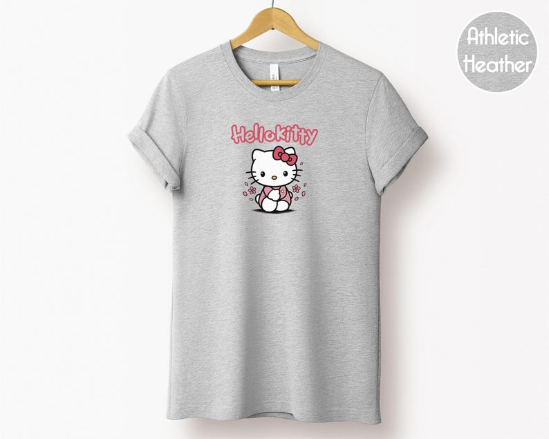 May include: A light gray t-shirt with a graphic of Hello Kitty. The design features Hello Kitty with a pink bow, surrounded by pink flowers, and the text "Hello Kitty" in pink. The shirt is hanging on a wooden hanger.
