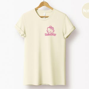 May include: Cream-colored t-shirt with a pink Hello Kitty graphic on the front. The graphic includes Hello Kitty's face and the text "Hello Kitty" in a cursive font. The shirt is hanging on a wooden hanger.