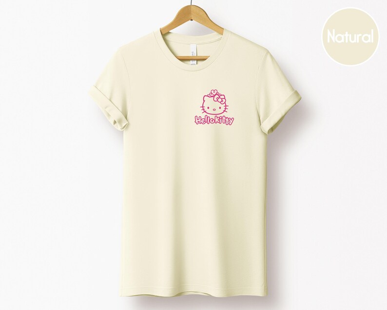 May include: Cream-colored t-shirt with a pink Hello Kitty graphic on the front. The graphic includes Hello Kitty's face and the text "Hello Kitty" in a cursive font. The shirt is hanging on a wooden hanger.
