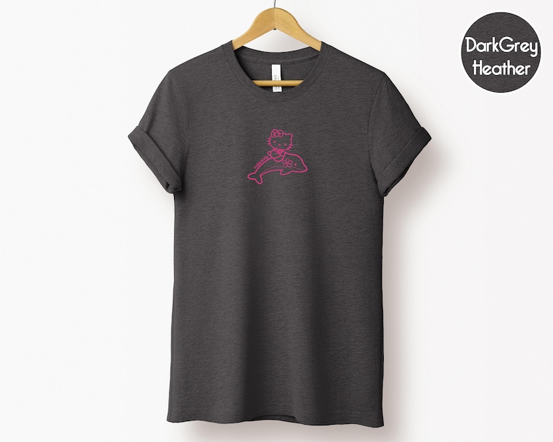 May include: Dark grey heather t-shirt with a pink outline graphic of a cartoon character riding a dolphin. The t-shirt is hanging on a wooden hanger against a white background. The text "Dark Grey Heather" is in a circle on the upper right.