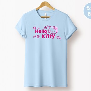May include: A pale blue t-shirt featuring the words "Hello Kitty" in pink, accompanied by a graphic of the character's face and floral accents. The t-shirt is displayed on a wooden hanger.