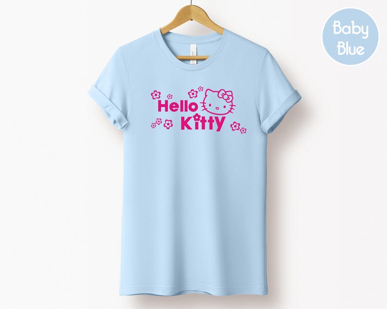 May include: A pale blue t-shirt featuring the words "Hello Kitty" in pink, accompanied by a graphic of the character's face and floral accents. The t-shirt is displayed on a wooden hanger.