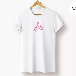 May include: White t-shirt with a pink graphic of a cartoon cat character riding a dolphin. The graphic is located on the front of the shirt. The t-shirt is hanging on a wooden hanger.