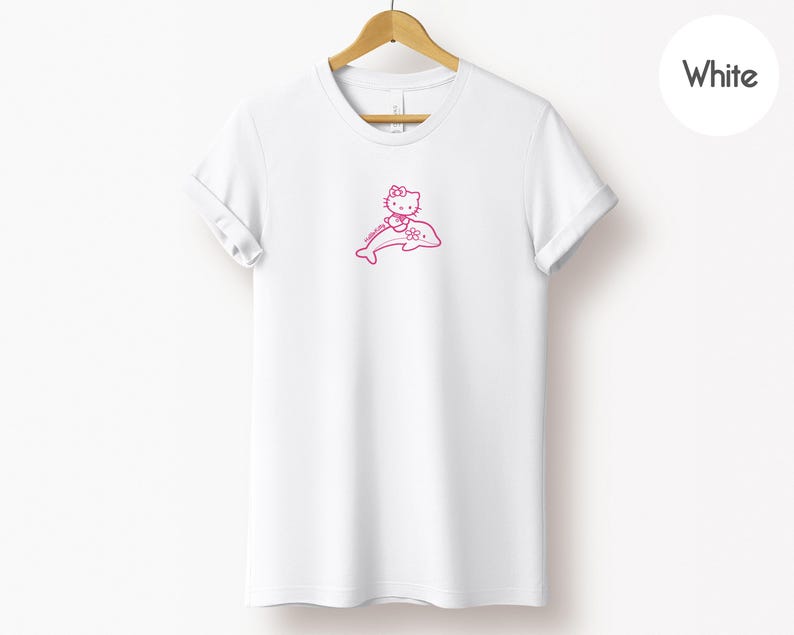 May include: White t-shirt with a pink graphic of a cartoon cat character riding a dolphin. The graphic is located on the front of the shirt. The t-shirt is hanging on a wooden hanger.