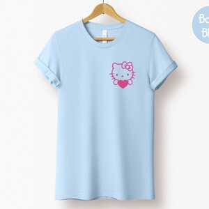 May include: A light blue t-shirt with rolled-up sleeves, hanging on a wooden hanger. The shirt features a pink graphic of a cartoon cat holding a heart. A circular "Baby Blue" label is in the upper right corner.