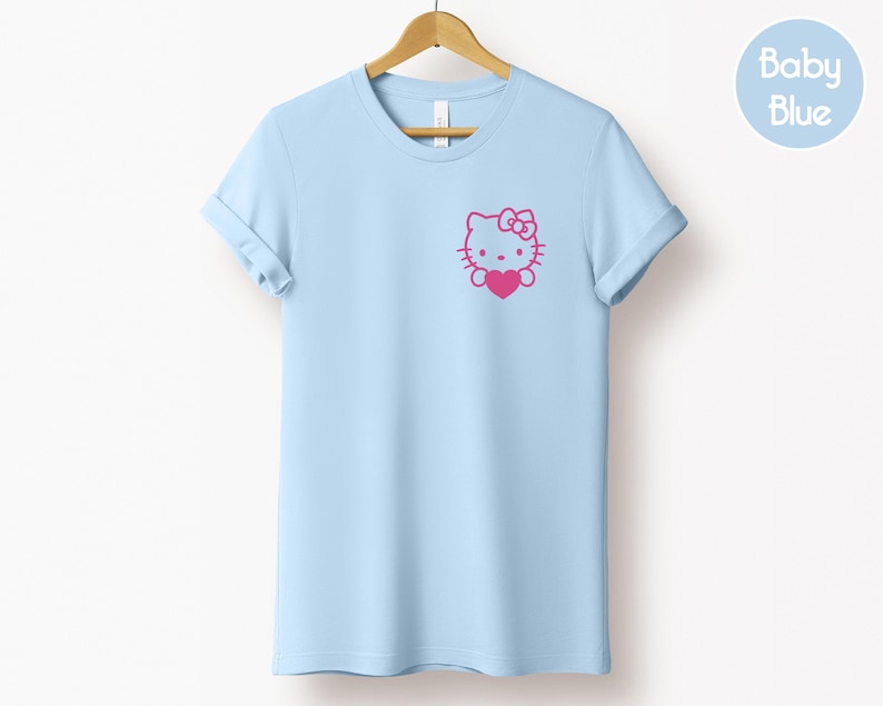 May include: A light blue t-shirt with rolled-up sleeves, hanging on a wooden hanger. The shirt features a pink graphic of a cartoon cat holding a heart. A circular "Baby Blue" label is in the upper right corner.