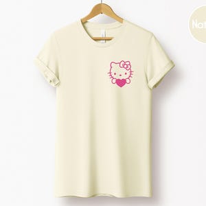 May include: Cream-colored t-shirt with a pink graphic of a cartoon cat holding a heart. The t-shirt is hanging on a wooden hanger. The graphic is on the left chest area of the shirt. The shirt has rolled-up sleeves.