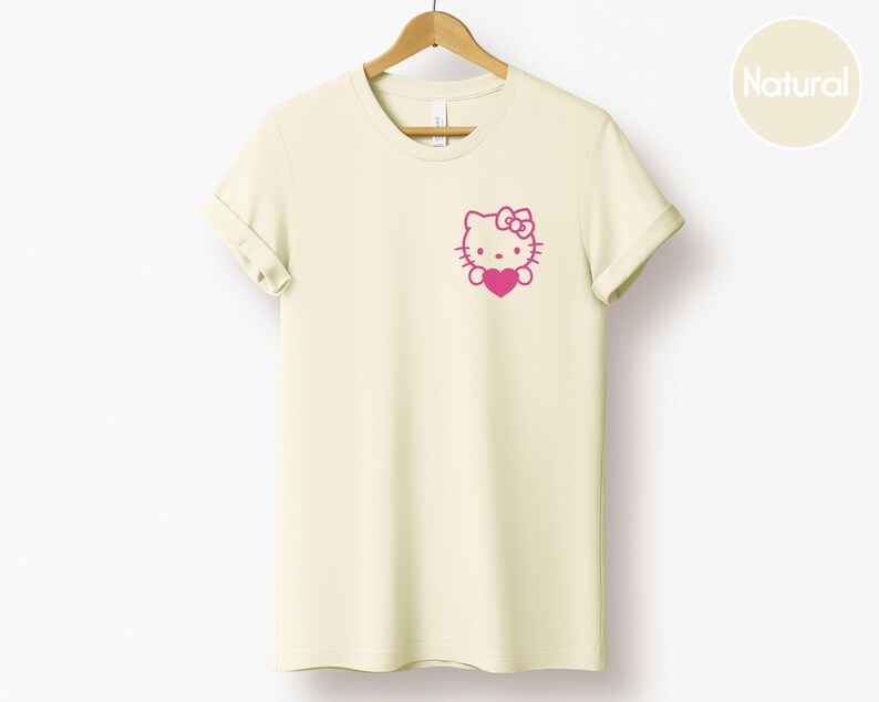 May include: Cream-colored t-shirt with a pink graphic of a cartoon cat holding a heart. The t-shirt is hanging on a wooden hanger. The graphic is on the left chest area of the shirt. The shirt has rolled-up sleeves.